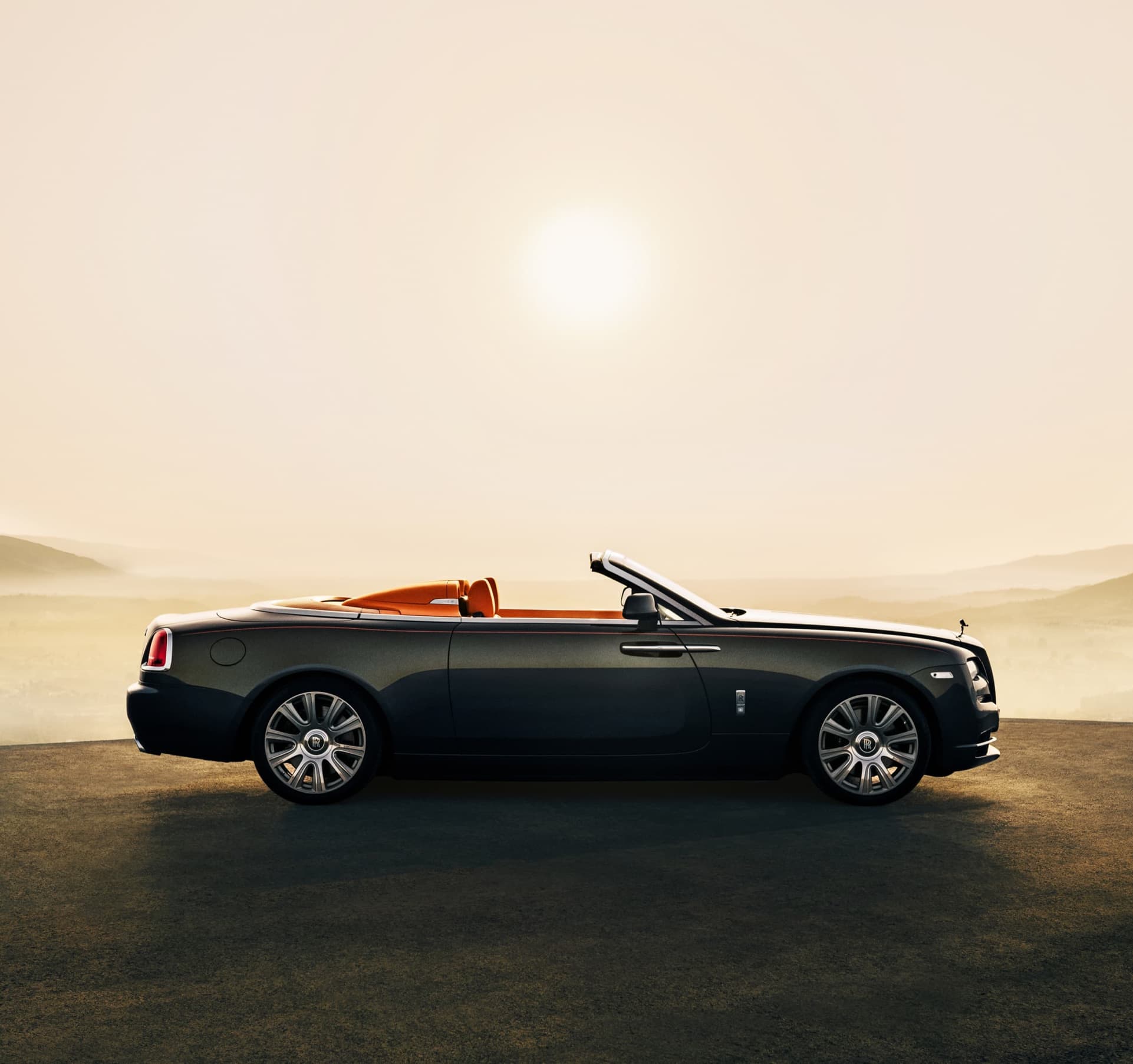 Side exterior of stationary Rolls-Royce Dawn motor car with Dawn Aero Cowling Tonneau Cover open on sunny hilltop with expansive views
