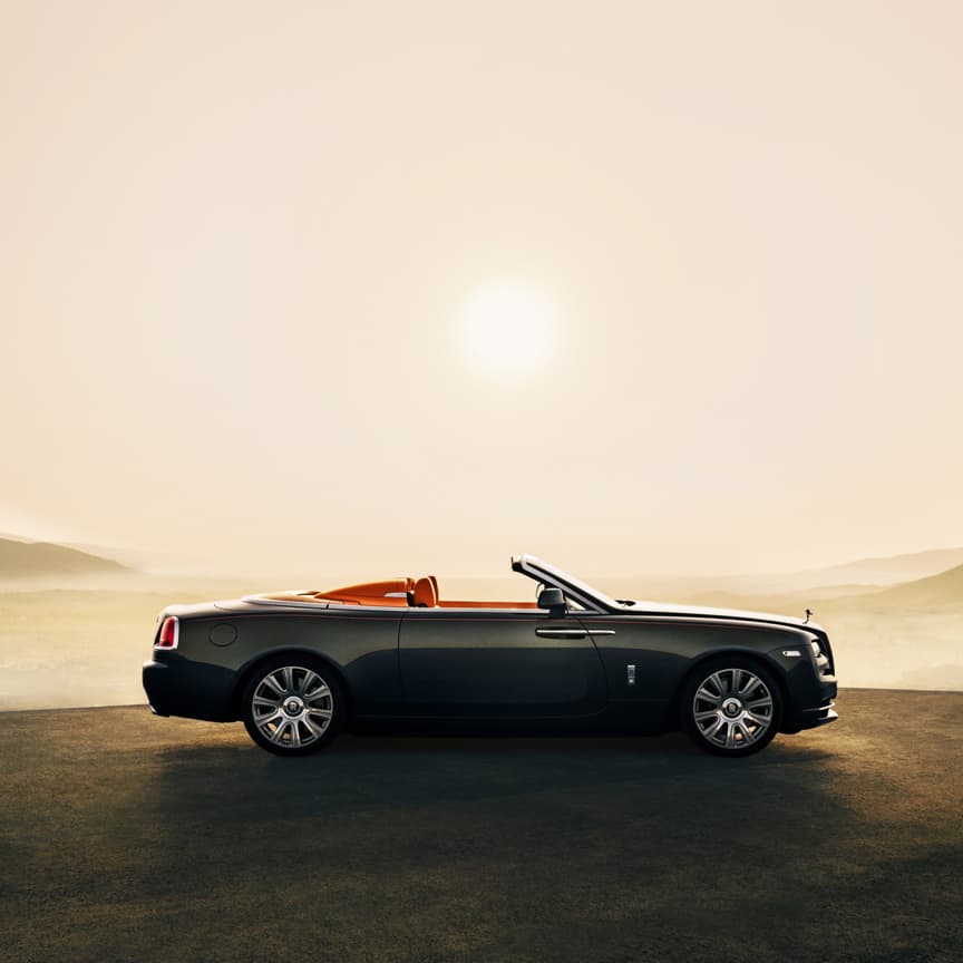 Side exterior of stationary Rolls-Royce Dawn motor car with Dawn Aero Cowling Tonneau Cover open on sunny hilltop with expansive views