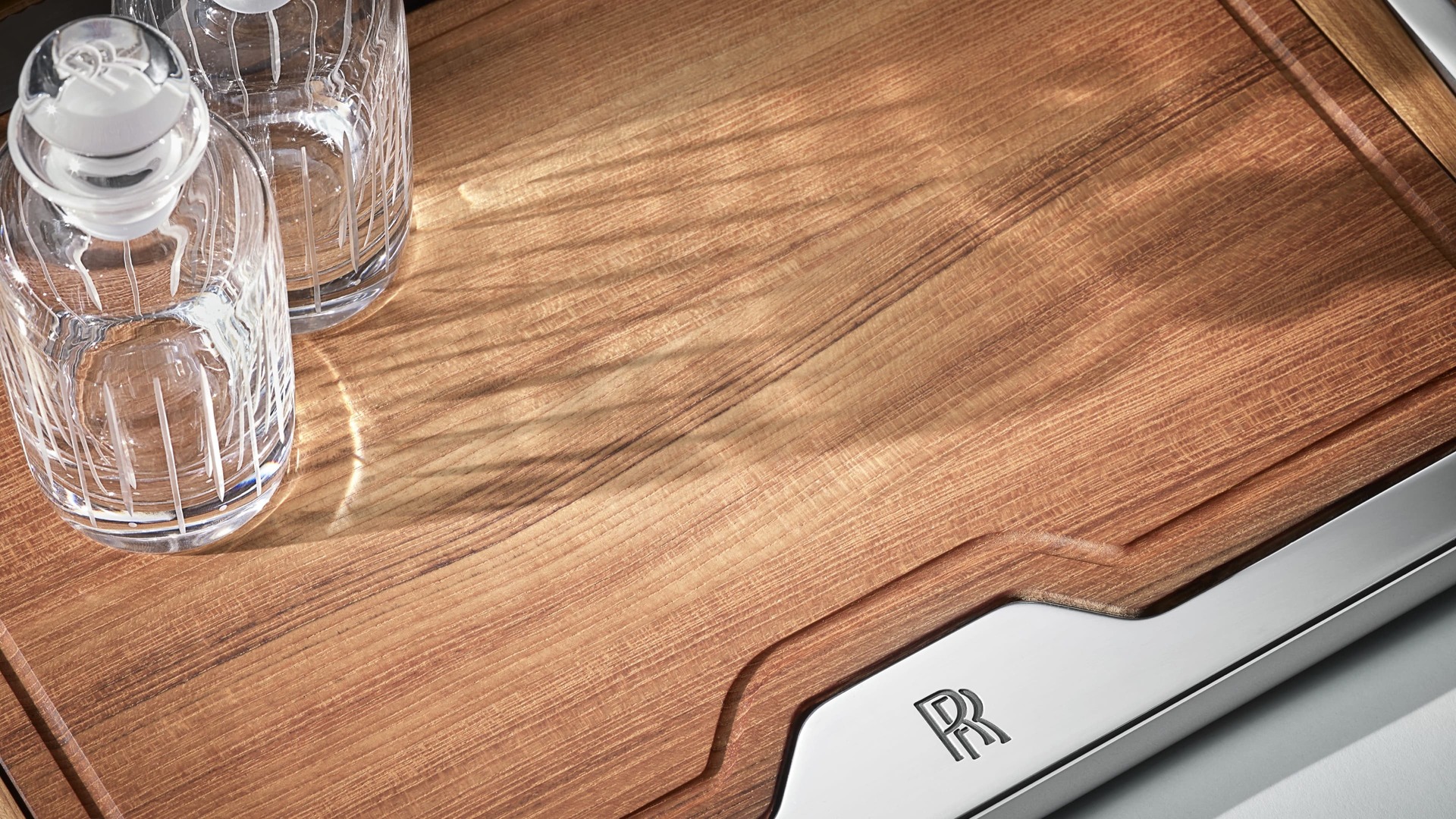 A close up shot of the wooden deck from the picnic hamper accessory, bordered with metal embossed with the Rolls-Royce monogram with two decanters placed ontop of the deck